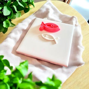 Puede incluir: Una baldosa cuadrada blanca con borde rosa, con un molde en forma de pez rosa y una impresión en forma de pez blanco. La baldosa descansa sobre una tela rosa claro, con follaje verde en primer plano y una superficie de madera.