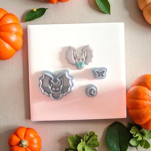 May include: A collection of gray plastic Halloween-themed cookie cutters, including bats, a butterfly, and a crescent moon, arranged on a white surface. Bright orange pumpkins and green leaves surround the cutters, creating a festive autumn scene.