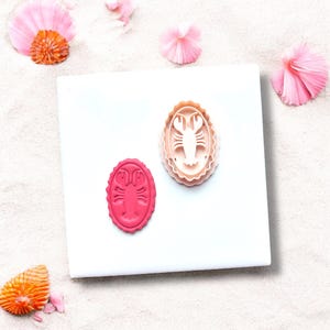 Könnte beinhalten: Zwei hummerförmige Ausstecher, einer rosa und einer beige, liegen auf einer weißen Oberfläche. Der rosa Ausstecher hat einen Hummerabdruck. Der beige Ausstecher ist ein dreiteiliges Set. Rosa und orangefarbene Muscheln sind verstreut.