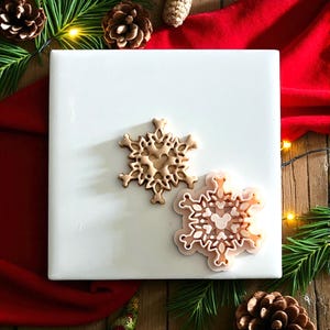 May include: Two snowflake-shaped cookie cutters. One is a light brown color, and the other is a light pink color with white accents. The cutters are on a white square surface, with pine needles, pine cones, and a red cloth in the background. Christmas-themed.