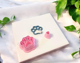 May include: Three cookie cutters in the shape of a paw print, a heart, and a paw print with a heart inside. The paw print cutters are pink and the heart cutter is blue.