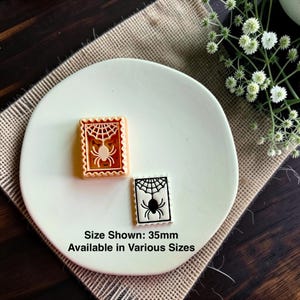 May include: A peach-colored rectangular cookie cutter and a white rectangular cookie with a black spider and web design. The text on the image states Size Shown: 35mm Available in Various Sizes. The items are displayed on a round white surface.