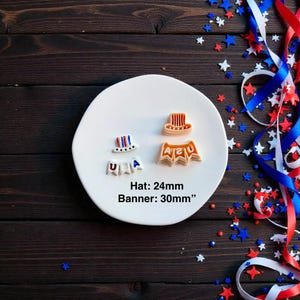 May include: A white plate displays patriotic-themed cookie cutters: a hat (24mm) and a banner (30mm) spelling "USA". Red, white, and blue stars and ribbons decorate the dark wood surface, creating a festive scene.