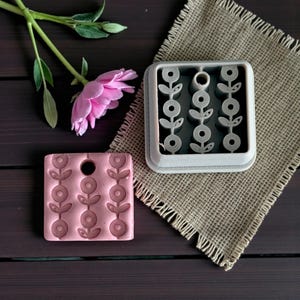 May include: A pink floral design stamp and a gray floral design mold sit on a dark wooden surface next to a pink flower and a burlap cloth. The stamp has a hole at the top. The mold is square.