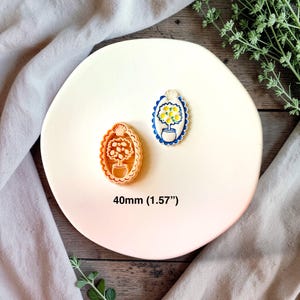 Puede incluir: Dos sellos de galletas ovalados con un diseño de árbol. Un sello es de color marrón claro, el otro azul, blanco y amarillo. Los sellos miden 40 mm (1,57") y están sobre un plato blanco.