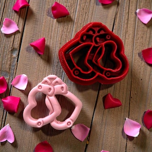 May include: A red heart-shaped cookie cutter and a pink heart-shaped object are displayed on a wooden surface, surrounded by scattered pink and red rose petals. The cookie cutter has a detailed design.
