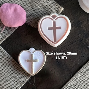 May include: Two heart-shaped pendants with a cross design. The pendants are white with a gold-colored outline and a cross in the center. The size shown is 28mm (1.10"). A pink petal is in the upper left corner.