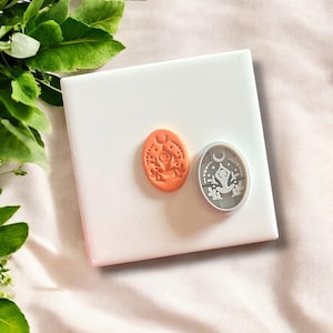 May include: Two oval-shaped stamps with nature-themed designs. One stamp is orange and features a floral design with a crescent moon. The other stamp is silver and has a frog in a yoga pose with a crescent moon and floral elements.