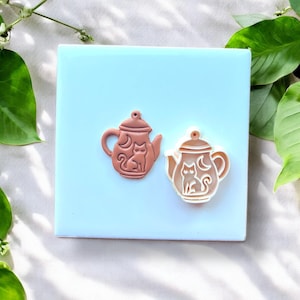 Op de afbeelding: Twee theepotvormige koekjessnijders. Eén is effen bruin, de andere is wit met een bruine omtrek. Elk heeft een kat en een halve maan ontwerp. De uitstekers liggen op een lichtblauwe vierkante tegel, omringd door groene bladeren.