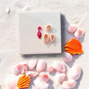 May include: A collection of pink and orange polymer clay cutters in the shape of a flamingo, leaves, and a seashell, arranged on a white square tile. The tile is surrounded by seashells and flower petals on a sandy surface.