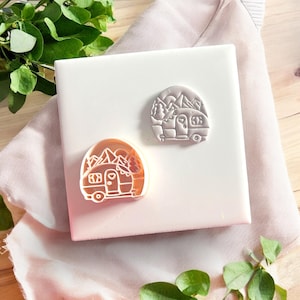 May include: Two camping-themed stamps. One stamp is orange and the other is white. Both stamps feature a camper with mountains and trees in the background. The stamps are on a white square surface with a pink gradient.