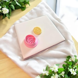 Puede incluir: Un juego de cortadores de galletas rosa y amarillo con un carácter chino para "doble felicidad" sobre una superficie blanca. El cortador se utiliza para crear galletas para una boda u otra ocasión especial.