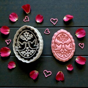 May include: Two cookie cutters with a dove design, one gray and one pink, are surrounded by pink heart shapes and rose petals on a dark wooden surface. The cookie cutters feature a tree and two doves.