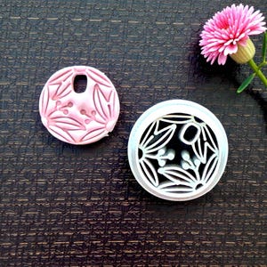 May include: Two circular, floral-patterned stamps. One is pink with a hole, the other is white with a black interior. A pink flower with a yellow center and green stem is in the background.