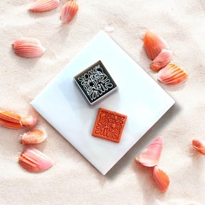 May include: A square stamp with a floral design, next to its impression. The stamp is in a black frame with a white design. The impression is in orange. The image is on a white square with seashells scattered around.