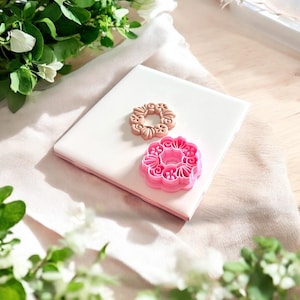 May include: Two cookie cutters, one brown and one pink, are on a white surface. The cutters are shaped like a wreath with intricate designs.