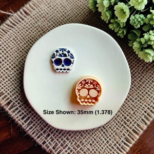 May include: Two skull-shaped items, one white with blue floral patterns and one light peach-colored plastic cutter, are displayed on a white circular surface. The text "Size Shown: 35mm (1.378)" is printed below the items, indicating their scale.