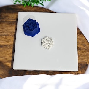 Puede incluir: Un molde de plástico azul de un dado de 20 caras y un dado de cerámica blanco de 20 caras sobre una baldosa blanca.