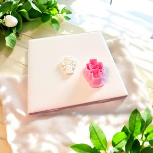 May include: Two cookie cutters, one white and one pink, shaped like a person wearing a dress. The cutters are on a white surface.