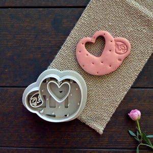 May include: A pink heart-shaped cookie and a matching cookie cutter sit on a burlap cloth. The cookie has a heart cutout and small dot details. The cutter is gray and has a heart and text design. A pink rose is in the corner.