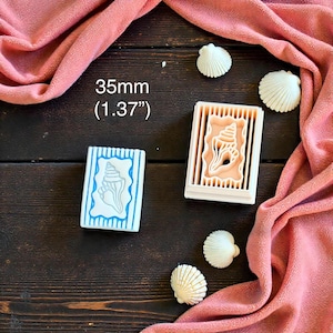 May include: Two seashell-themed cookie cutters and three seashell decorations are displayed on a dark wooden surface, with a pink fabric draped around them. One cutter is white with a blue seashell design, and the other is a beige seashell-shaped cutter. The text "35mm (1.37")" is visible.