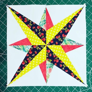 May include: A square quilt block featuring a vibrant star design. The star's points alternate between yellow polka dots, navy blue fabric with red strawberries, and red fabric. The background is white, and the block is on a green cutting mat.