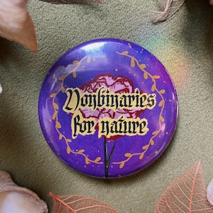 May include: A circular purple badge featuring the text "Nonbinaries for nature" in gold lettering. A floral design is in the center, with gold vines around the edge. The badge is surrounded by dried leaves.