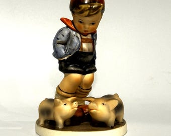 May include: A ceramic figurine of a young boy in a blue jacket, black overalls, and an orange cap, gazing at two small pigs. The figurine is set on a round base with a brown and beige colour scheme.