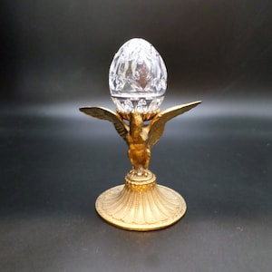 May include: A decorative egg-shaped crystal object sits atop a gold-colored metal stand. The stand features an eagle design with outstretched wings and a detailed base. The crystal egg has a cut-glass pattern.