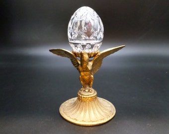 May include: A decorative egg-shaped crystal object sits atop a gold-colored metal stand. The stand features an eagle design with outstretched wings and a detailed base. The crystal egg has a cut-glass pattern.