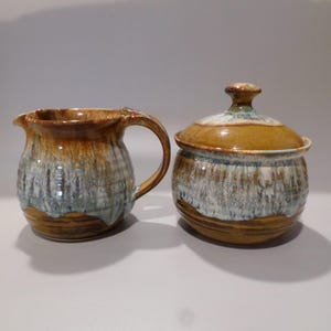 Könnte beinhalten: Ein braunes und weißes Keramik-Sahnegießer- und Zuckergefäß-Set mit glasierter Oberfläche. Der Sahnegießer hat einen Griff und das Zuckergefäß einen Deckel.