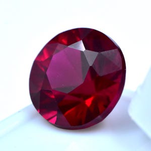 Pode incluir: Uma gema de rubi facetada, em forma oval. A gema é de uma cor vermelha profunda, com múltiplas facetas que refletem a luz. A gema está colocada sobre um fundo branco, realçando o seu brilho e clareza.