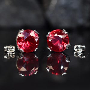 May include: A pair of stud earrings featuring large, round, faceted red gemstones set in silver-colored metal. The earrings are displayed on a reflective black surface, with the earring backs visible.