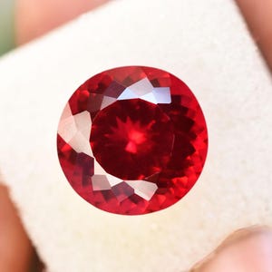 Può includere: Una gemma sfaccettata, taglio rotondo, di colore rosso intenso. La gemma è tenuta su uno sfondo bianco, mostrando le sue brillanti sfaccettature e i riflessi interni. Il colore della gemma è un rosso ricco e saturo.