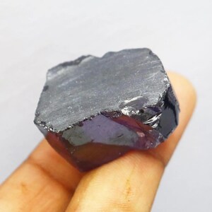 Puede incluir: Una gema en bruto de color p&uacute;rpura oscuro, con una superficie rugosa y texturizada. El cristal se sostiene en una mano, mostrando su forma natural e irregular y su color profundo. La piedra parece tener un ligero brillo.