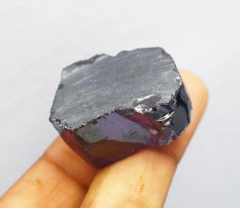 Puede incluir: Una gema en bruto de color p&uacute;rpura oscuro, con una superficie rugosa y texturizada. El cristal se sostiene en una mano, mostrando su forma natural e irregular y su color profundo. La piedra parece tener un ligero brillo.