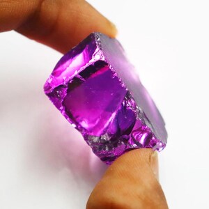 Puede incluir: Una vibrante gema de amatista en bruto, de un tono p&uacute;rpura intenso. Los bordes irregulares del cristal reflejan la luz, mostrando su textura natural y variaciones de color. Esta gema es un hermoso ejemplo de formaciones minerales naturales.