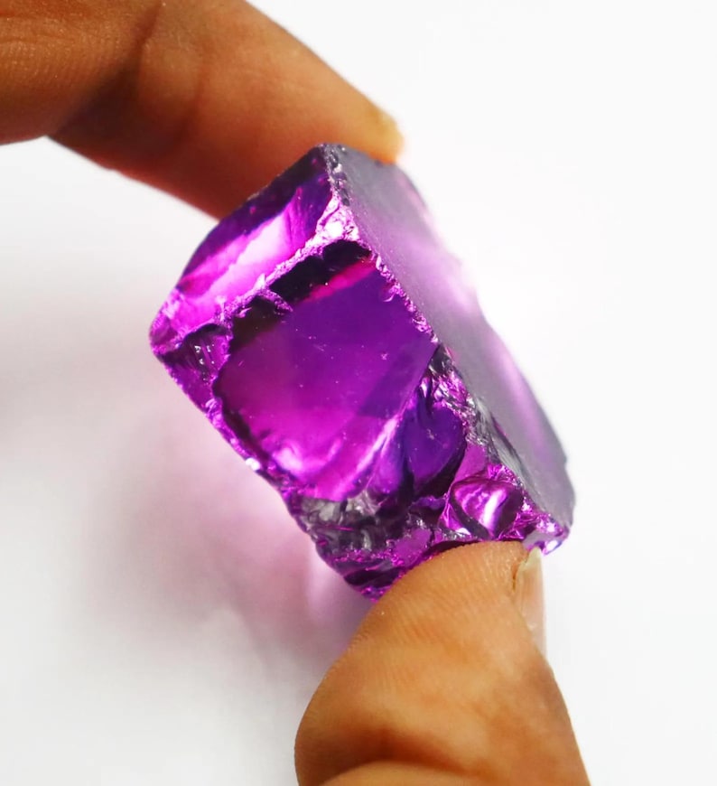 Puede incluir: Una vibrante gema de amatista en bruto, de un tono p&uacute;rpura intenso. Los bordes irregulares del cristal reflejan la luz, mostrando su textura natural y variaciones de color. Esta gema es un hermoso ejemplo de formaciones minerales naturales.