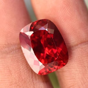 Può includere: Una gemma sfaccettata, taglio a cuscino, di colore rosso intenso. La superficie della gemma riflette la luce, mostrando i suoi intricati tagli e la sua brillantezza interna. La pietra è tenuta in una mano.
