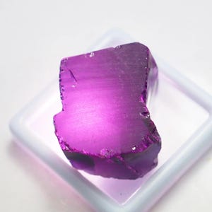 Puede incluir: Una vibrante gema de amatista en bruto, de un tono p&uacute;rpura intenso. La piedra en bruto se exhibe en un recipiente cuadrado blanco, mostrando sus bordes naturales e irregulares y su estructura cristalina. La gema est&aacute; iluminada, resaltando su color y textura.
