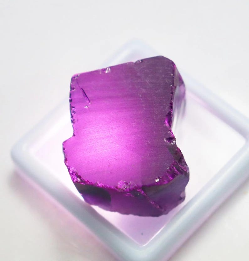 Puede incluir: Una vibrante gema de amatista en bruto, de un tono p&uacute;rpura intenso. La piedra en bruto se exhibe en un recipiente cuadrado blanco, mostrando sus bordes naturales e irregulares y su estructura cristalina. La gema est&aacute; iluminada, resaltando su color y textura.