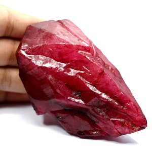 May include: A large, rough, red ruby crystal with a pointed tip. The crystal has a deep, rich red colour and is translucent.