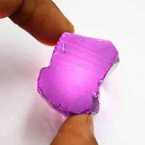 Puede incluir: Una gema p&uacute;rpura pulida de forma irregular. La gema se sostiene entre dos dedos, mostrando su superficie lisa y reflectante y su rico color. La piedra parece ser un corte en bruto, con bordes naturales visibles.