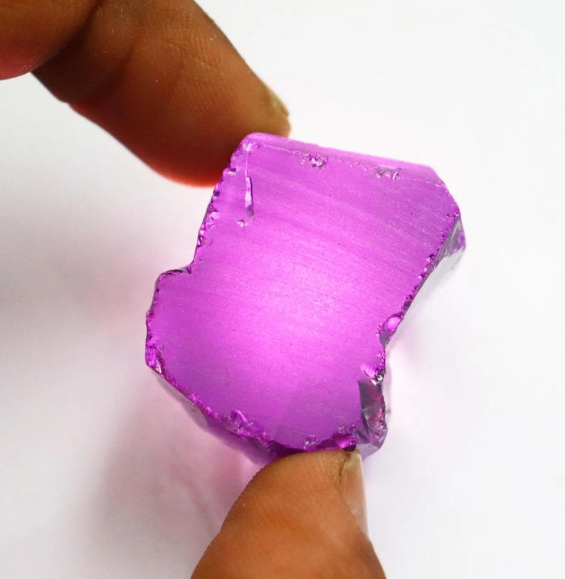 Puede incluir: Una gema p&uacute;rpura pulida de forma irregular. La gema se sostiene entre dos dedos, mostrando su superficie lisa y reflectante y su rico color. La piedra parece ser un corte en bruto, con bordes naturales visibles.