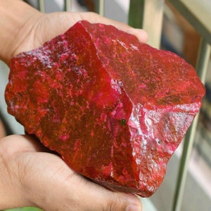 Könnte beinhalten: Ein großer, roher, roter Rubin-Edelstein. Der Rubin hat eine tiefrote Farbe mit einem leichten Orangestich. Der Edelstein ist unregelmäßig geformt und hat eine glatte, polierte Oberfläche.