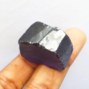 Puede incluir: Una gema en bruto de color p&uacute;rpura oscuro con una superficie cristalina reflectante. La piedra en bruto, de forma rectangular, se sostiene en una mano sobre un fondo blanco. La gema parece ser un esp&eacute;cimen mineral natural.