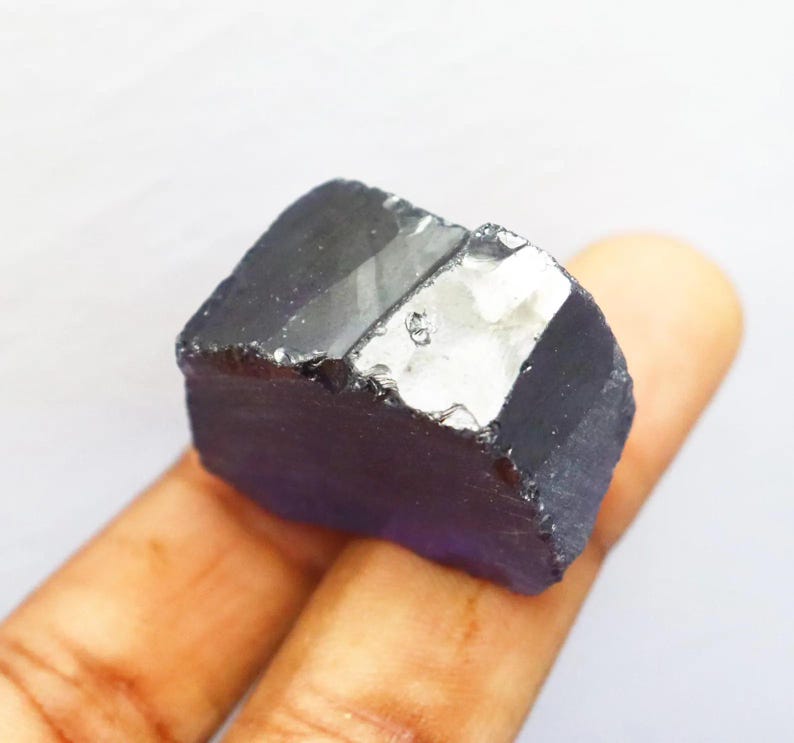 Puede incluir: Una gema en bruto de color p&uacute;rpura oscuro con una superficie cristalina reflectante. La piedra en bruto, de forma rectangular, se sostiene en una mano sobre un fondo blanco. La gema parece ser un esp&eacute;cimen mineral natural.