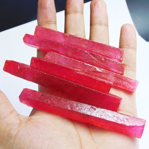 May include: A collection of six raw ruby crystals, each a long, slender, rectangular shape, with a deep red color and a slightly translucent appearance. The crystals are arranged in a pile on a hand.