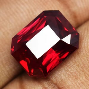 May include: A deep red gemstone with a rectangular emerald cut. The gemstone is faceted and has a high polish.