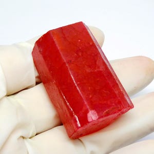 May include: A faceted, rectangular, red gemstone with a glossy finish. The gemstone is held in a gloved hand, showcasing its vibrant color and geometric shape. The stone's facets catch the light, highlighting its crystalline structure.